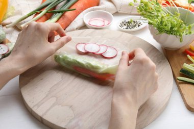 Woman wrapping spring roll at white wooden table with products, closeup