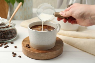 Woman pouring milk into cup with coffee at white wooden table, closeup