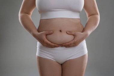 Gri arka planda göbek yağına dokunan kadın, yakın plan. Aşırı kilolu