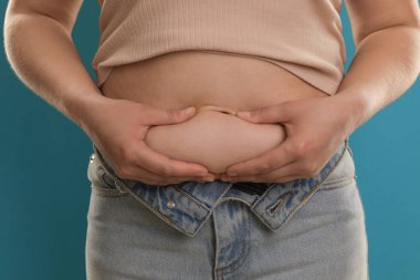 Açık mavi arka planda göbek yağına dokunan kadın, yakın plan. Aşırı kilolu