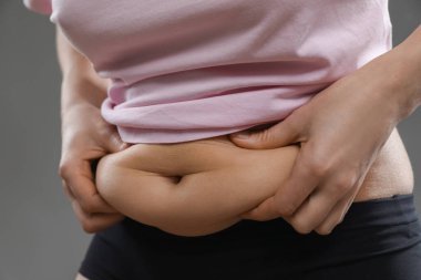 Gri arka planda göbek yağına dokunan kadın, yakın plan. Aşırı kilolu