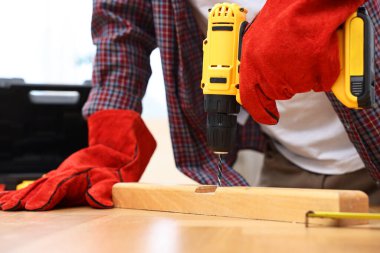 Young handyman working with electric drill at table in workshop, closeup