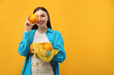 Woman holding string bag of fresh oranges and covering eye with fruit on orange background, space for text