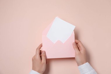 Woman holding letter envelope with card at beige table, top view