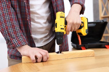 Young handyman working with electric drill at table in workshop, closeup
