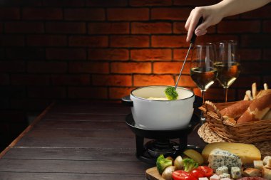 Woman dipping piece of broccoli into fondue pot with melted cheese at wooden table with wine and snacks, closeup. Space for text