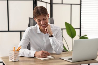 İçerideki ahşap masada giysi giyerken not alan adam.