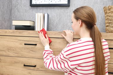 Woman cleaning wooden chest od drawers with rag at home