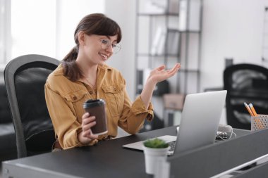 Woman with cup of coffee watching webinar at table in office
