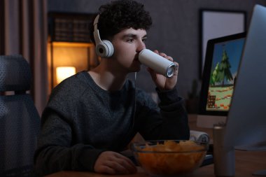 Young man with energy drink and headphones playing video game at wooden desk indoors