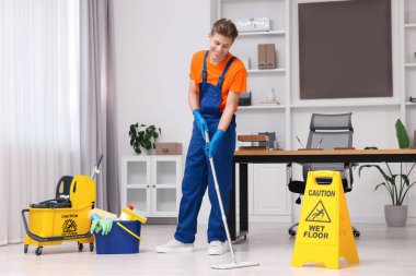 Temizlik işçisi yerleri paspasla yıkıyor. Ofiste içi malzeme dolu kova ve ıslak tabela var.