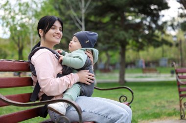 Anne çocuğunu parkta bankta askında (bebek taşıyıcısı) tutuyor. Metin için boşluk