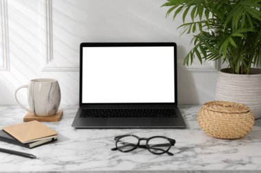 Beyaz duvarın yanındaki mermer masada bilgisayar, gözlük, bardak ve kırtasiye malzemesi olan bir ofis.