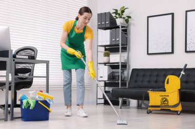 Temizlik işçisi yerleri paspasla yıkıyor. Ofiste içi malzeme dolu kova ve ıslak tabela var.