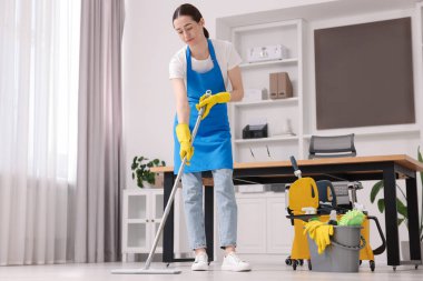 Temizlik işçisi yerleri paspasla yıkıyor, düşük açılı. Ofiste içi malzeme dolu bir kova.