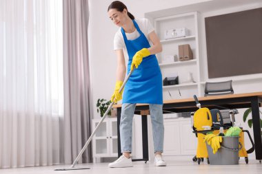 Temizlik işçisi yerleri paspasla yıkıyor, düşük açılı. Ofiste içi malzeme dolu bir kova.