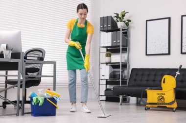 Temizlik işçisi yerleri paspasla yıkıyor. Ofiste içi malzeme dolu kova ve ıslak tabela var.