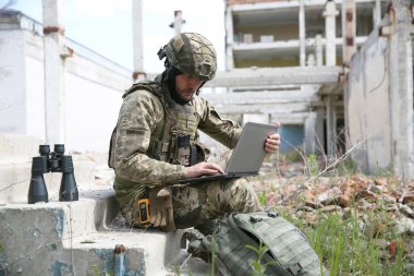 Askeri görev. Dışarıdaki terk edilmiş binanın yanında dizüstü bilgisayar kullanan üniformalı bir asker.