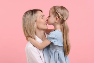 Daughter hugging and kissing her happy mother on pink background