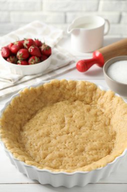 Kısa hamur işi yapıyorum. Hamur hamuru fırın tabağında, çilek, şeker, merdane ve süt beyaz ahşap masada.