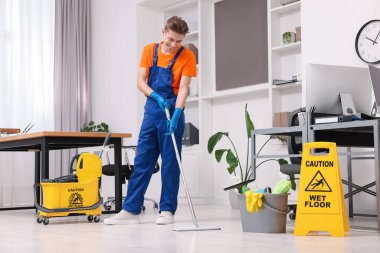 Temizlik işçisi yerleri paspasla yıkıyor. Ofiste içi malzeme dolu kova ve ıslak tabela var.