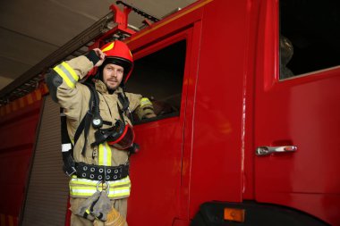 İstasyondaki kırmızı itfaiye aracının yanındaki üniformalı itfaiyecinin portresi.