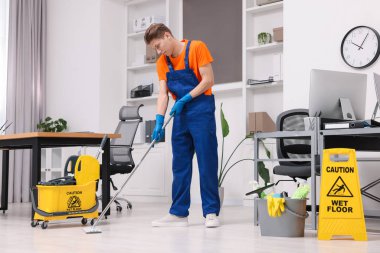 Temizlik işçisi yerleri paspasla yıkıyor. Ofiste içi malzeme dolu kova ve ıslak tabela var.