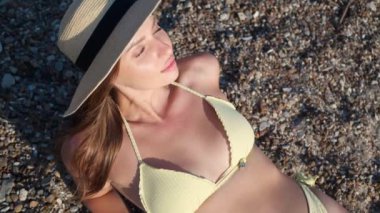Şapkalı güzel bir kadın çakıl taşı plajında güneşleniyor, manzaranın üstünde. Kamera aşağı iniyor