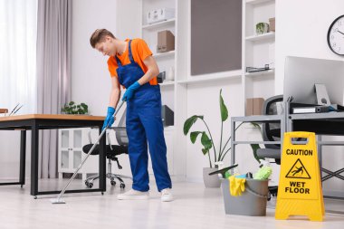 Temizlik işçisi yerleri paspasla yıkıyor. Ofiste içi malzeme dolu kova ve ıslak tabela var.