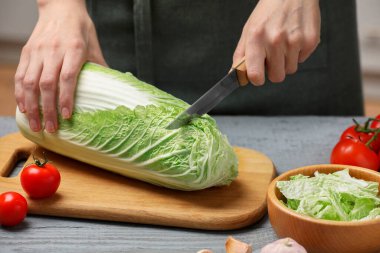 Woman cutting fresh chinese cabbage at grey wooden table in kitchen, closeup