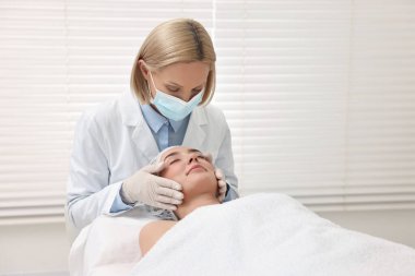 Tıbbi maskeli bir dermatolog klinikte hastanın yüzünü muayene ediyor.