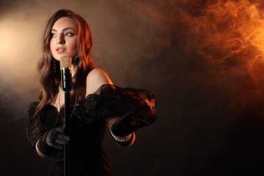 Beautiful young woman with microphone singing in color lights and smoke, space for text