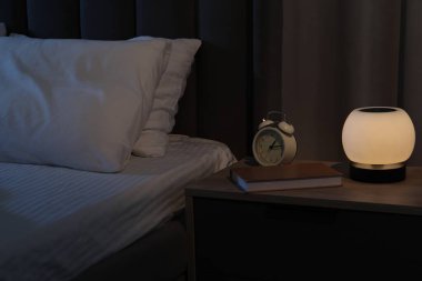 Gece lambası, çalar saat ve kitap yatağın yanındaki komodinin üzerinde.