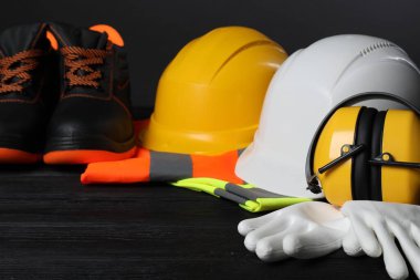 Bir çift çalışma botu, koruyucu şapkalar, koruyucu eldivenler, kulaklık ve siyah ahşap yüzeyde yansıtıcı yelekler, yakın plan.