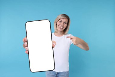 Açık mavi arka planda boş ekranlı cep telefonunu gösteren mutlu kadın. Tasarım için model