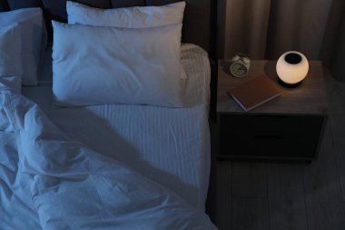 Gece lambası, çalar saat ve kitap yatağın yanındaki masanın üstünde, içeride, manzaranın üstünde.