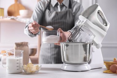 Kadın, kapalı mekânda hamur toplarken tezgah karıştırıcısına şeker ekliyor.