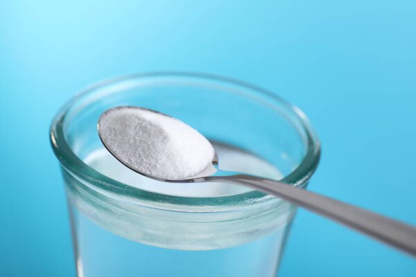 Ложка с пищевой содой над стаканом воды на светло-голубом фоне, крупный план