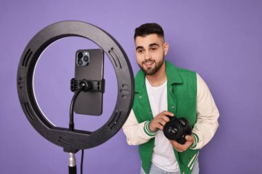 Teknoloji blogcusu kamerayı inceliyor ve mor arka planda akıllı telefon ve halka lambasıyla video kaydediyor.