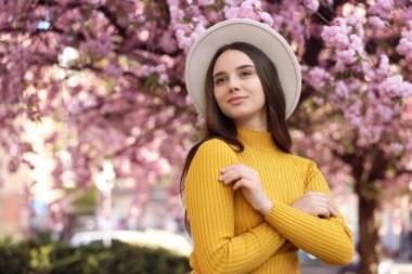 Beautiful woman in hat near blossoming tree on spring day, space for text