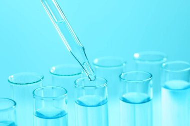 Açık mavi arka planda pipetten test tüpüne sıvı damlatılıyor, yakın plan. Laboratuvar analizi