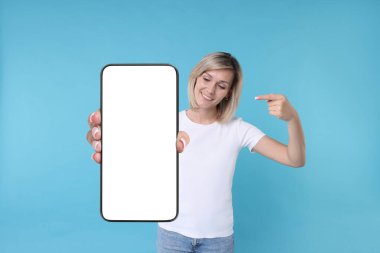 Açık mavi arka planda boş ekranlı cep telefonunu gösteren mutlu kadın. Tasarım için model