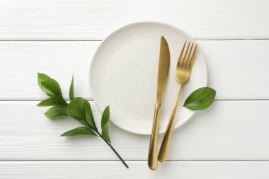 Çatal bıçak takımı, yeşil yapraklar ve beyaz ahşap masa üzerindeki tabaklar düz.
