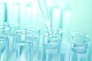 Turkuaz arka planda pipetten test tüpüne sıvı damlatılıyor.