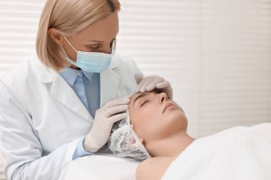 Tıbbi maskeli bir dermatolog klinikte hastanın yüzünü muayene ediyor.