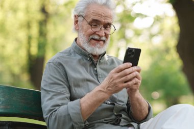 Parktaki bankta akıllı telefon kullanan mutlu büyükbabanın portresi.