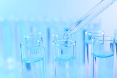 Açık mavi arkaplanda pipetten test tüpüne sıvı damlatılıyor, yakın plan.