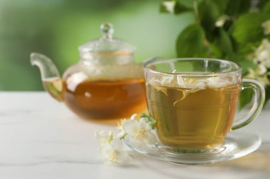 Aromatik yasemin çayı fincanda, çiçekler, çaydanlık ve yeşil yapraklar beyaz mermer masada.