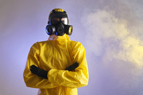 Worker wearing gas mask in smoke on violet background, low angle view. Space for text