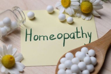 Üzerinde kelime olan yapışkan bir not Homeopati, haplar ve ahşap masa üzerindeki papatya çiçekleri, yakın plan.